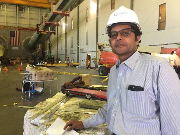 A man in a warehouse wears a white helmet leans against a small surface that is holding long metal rulers and other equipment.