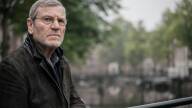 A man with grey hair and stubble stands before a blurry waterway.