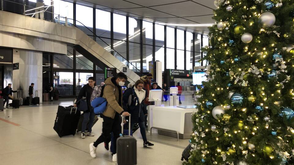 People with suitcases walk through an airport terminal. A Christmas tree decorated with ornaments is visible in the foreground.