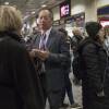 A man in a gray suit gestures with his hands as he speaks to a woman standing in front of him in a large train station, surrounded by other people.