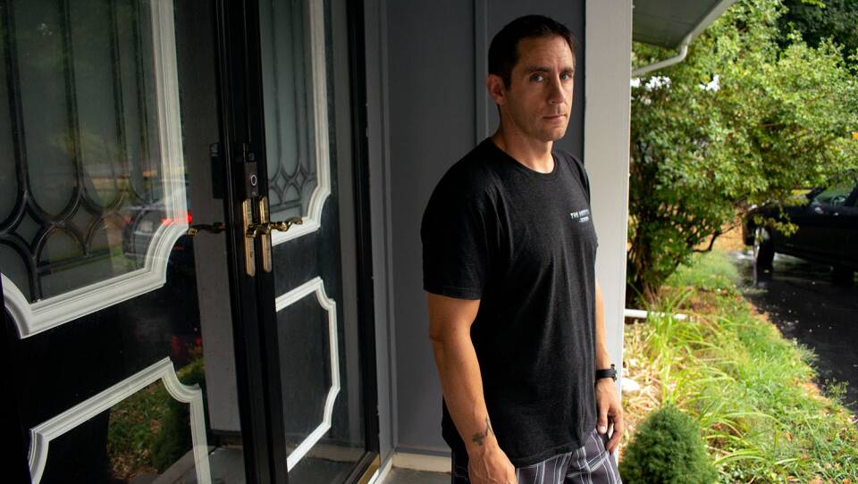 A man in a T shirt stands on the front porch of a home. He looks concerned and is resting his arms at his sides.