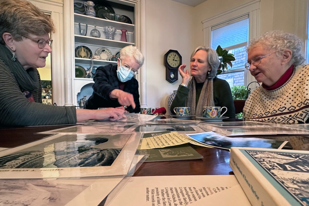 Four older women sit around a wooden table strewn with documents, drinking tea. They're debating and digging through the papers.