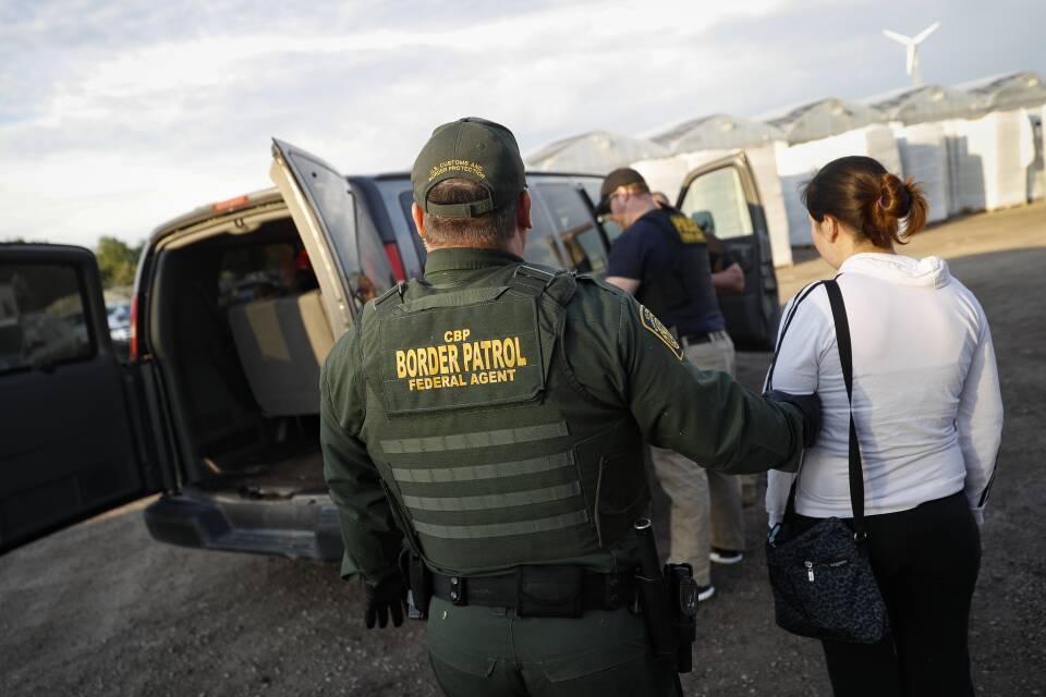 A person wearing a CBP Border Patrol Federal Agent uniform holds a woman by the arm as they lead her to a van where a couple other people are.