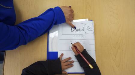 A young boy slowly writes "Daniel" with a pencil, as a teacher's hand points letter by letter.