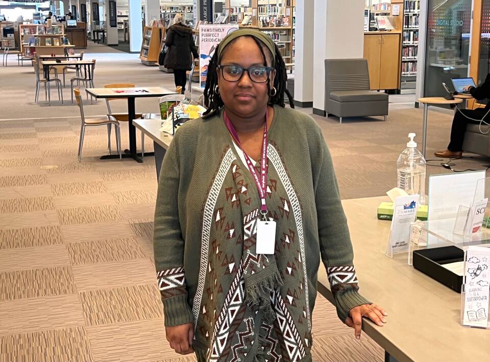 A woman with an ID badge hanging from a lanyard around her neck stands in the middle of an open library area.