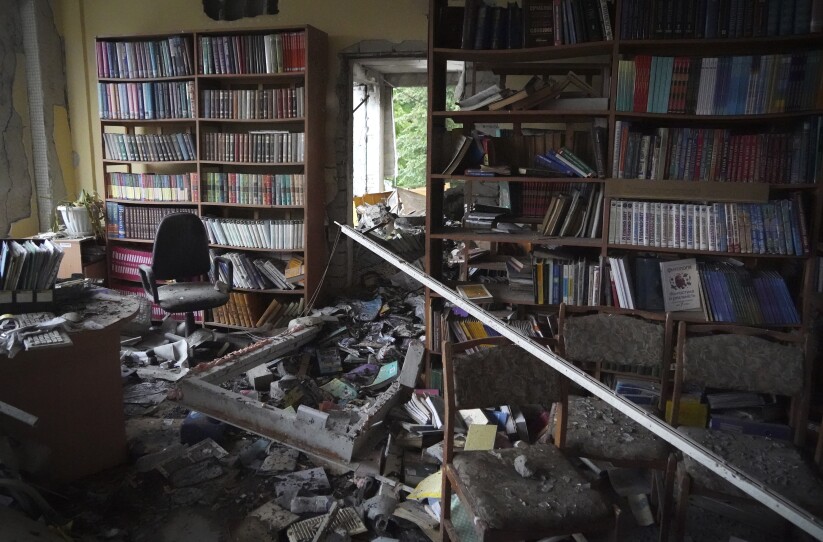 Rubble and dust cover a small library room. The door has been blown to pieces, and a tree outdoors is visible through the opening.