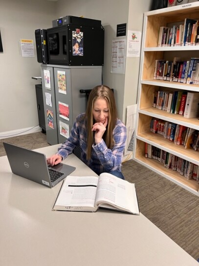 A women in a blue plaid shirt and long blonde hair sits at a desk with a laptop and open textbook.