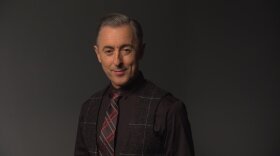 This is a headshot of a man. He is wearing a dark grey  plaid blaze, a black shirt and a grey and red argyle patterned tie.  He is standing against a dark grey background, he has a bemused expression on his face, smiling slightly.