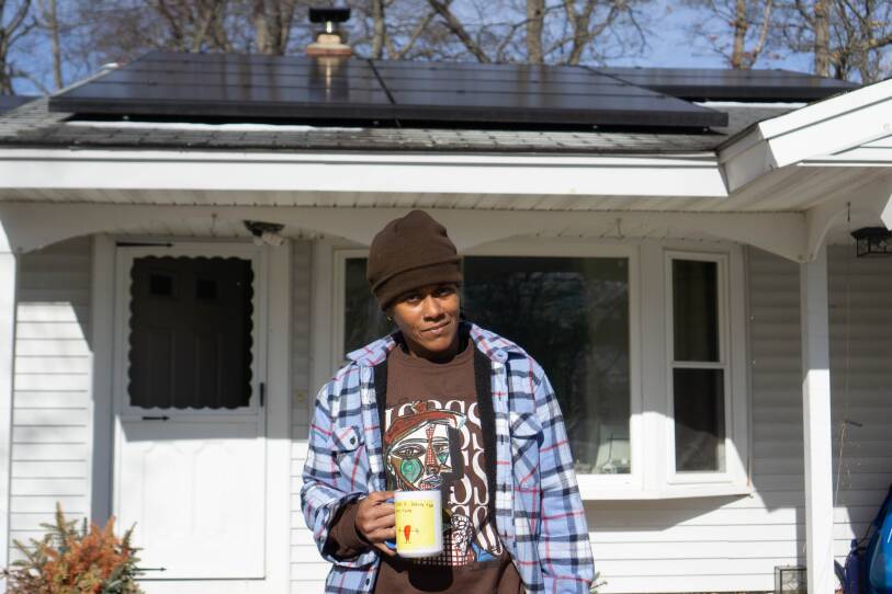 A woman in a brown hat and plaid shirt stands in front of a white house with solar panels on the roof.