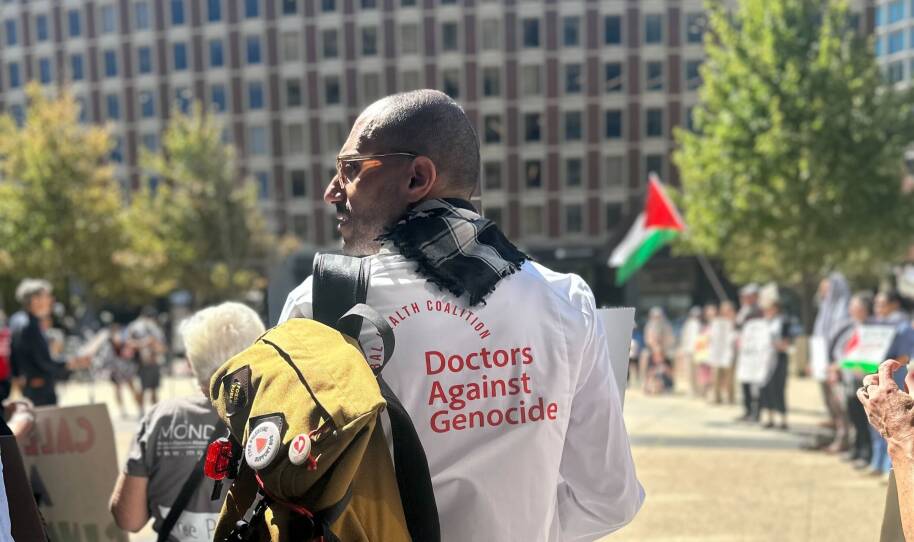 A man with his back to the camera wearing a shirt that says Doctors Against Genocide. In the background is another person waving a Palestinian flag.