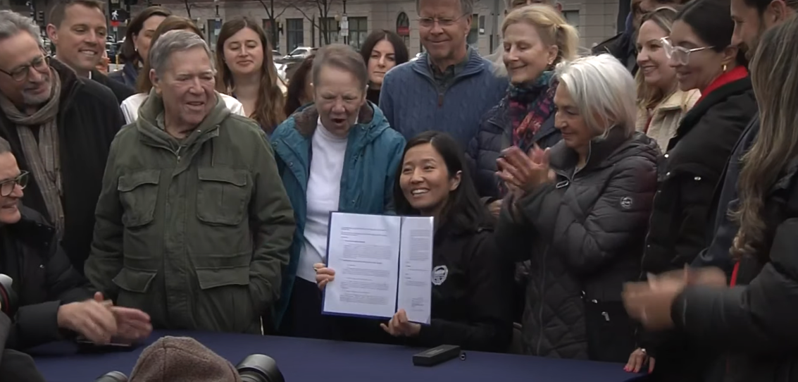 A woman holds up a piece of legislation, surrounded by more than a dozen enthusiastic watchers.