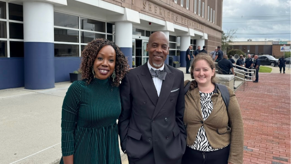 Two women and a man pose for a photo in front of the Suffolk County Sheriff's Department with a cluster of uniformed officers sitting and relaxing in the background.
