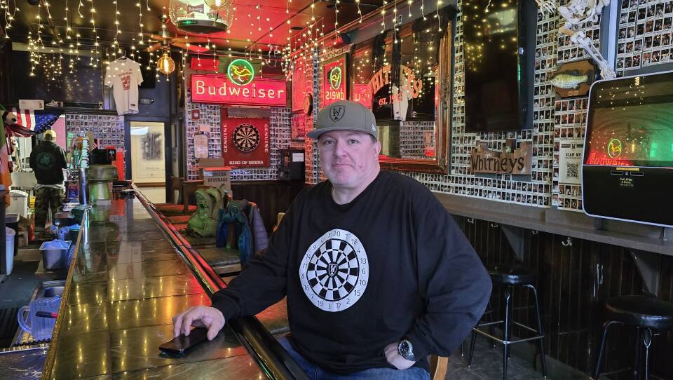 Man in a green cap and blue sweatshirt stands behind a bar with many framed pictures, hanging lights and other paraphernalia.
