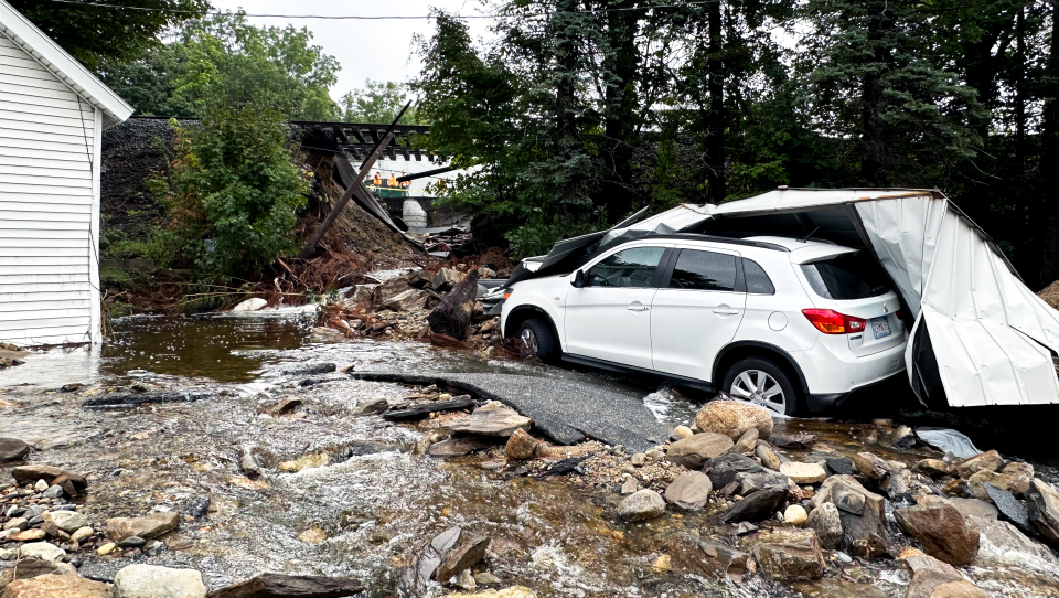 Water is still flooding an area — a car is under a broken aluminum shed that looks to have been partly carried away by flooding. In the background, a train track is suspended over nothing.