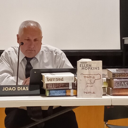 A man in a collared shirt and tie sits behind a table, looking at a laptop computer, as stacks of books are displayed in front of him.