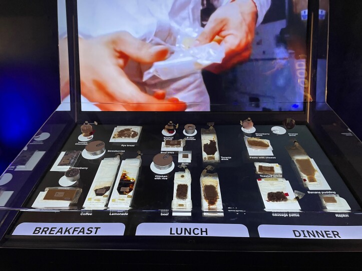 This is a photograph of a display case, which contains dehydrated food from space missions. Though the case is organized by breakfast, lunch and dinner, all the foods look the same, small brown squares. The food labeled "sausage pattie" looks no different than the packet of dehydrated coffee.
