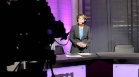 The governor stands on GBH's purple set in a radio headset and blazer, listening intently. She's facing a camera that stands in the foreground, obscured in the shadows.