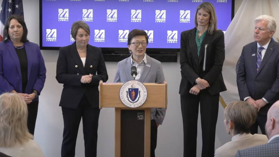 A woman with short dark hair and glasses stands at a lectern bearing the state seal of Massachusetts, in front of a blue background bearing the UMass Lowell logo.