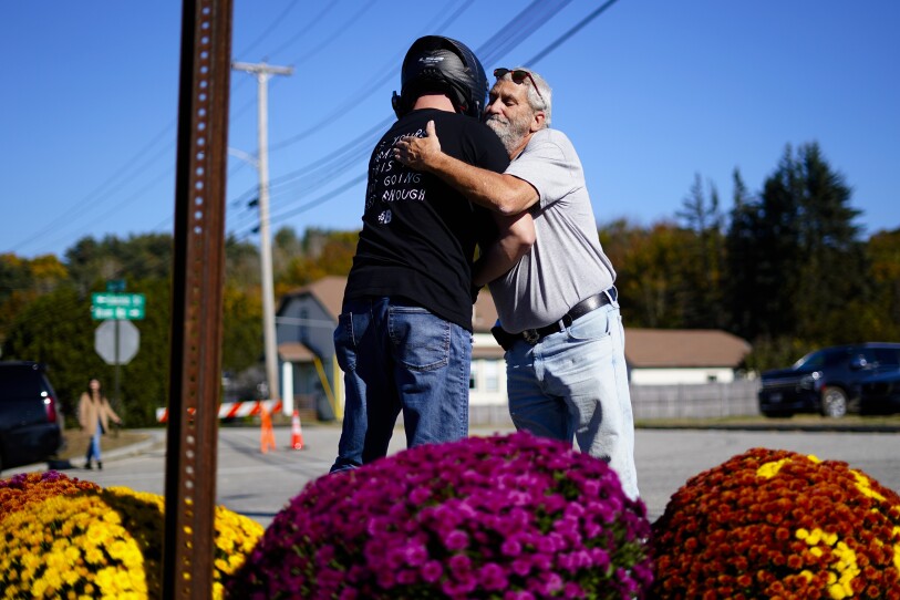 A man hugs a person wearing a motorcycle helmet in front of large mum displays on a city street.