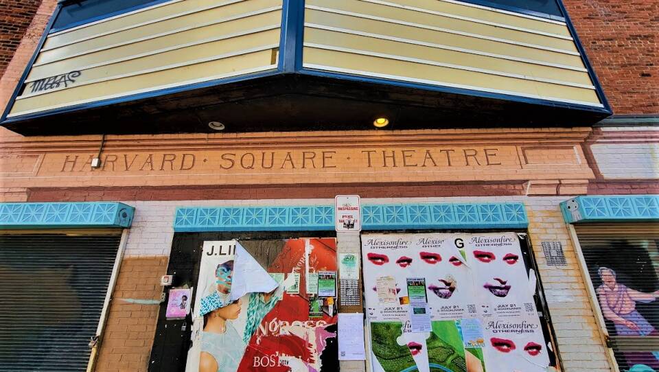 An assortment of posters, some tattered and torn, line the doors of a building with the words "Harvard Square Theatre" on a sign.