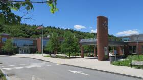 A sign reading "Wayland High School" and a clock mark the entrance to a large brick and glass building.