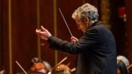 A man with gray hair and glasses conducts an orchestra.