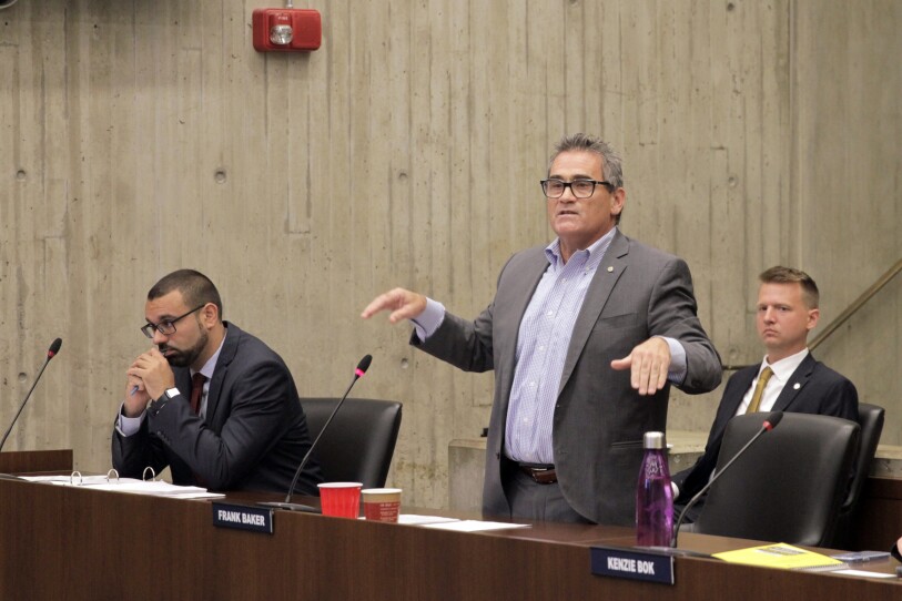 A man speaks emphatically, motioning with his hands, while the man seated next to him is hunched over and looking off into the distance