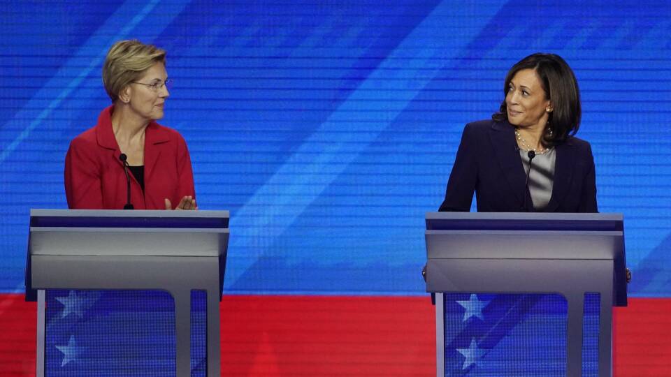 Two women stand at podiums in front of a blue-and-red backdrop.