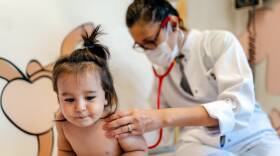 An adult in a white coat sits on an exam table behind a toddler, placing their stethoscope on the child's back while holding them still.