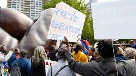 A protester holds a sign reading "Needed: justice, equity, education, outreach, voice" as they stand in a crowd in a park.