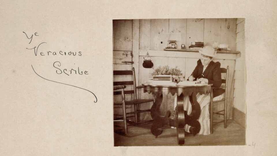 A photograph is pasted into the journal, captioned in handwriting "Ye Veracious Scribe." The photo shows a woman in 19th-century clothes sitting at a wooden table, writing. 