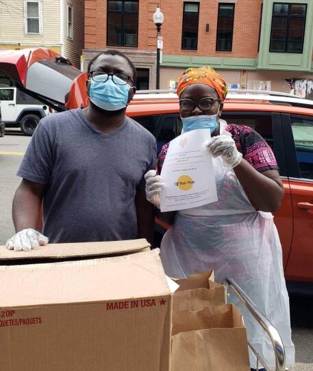 Two people wearing medical masks stand behind cardboard boxes and brown paper bags.