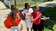 Sisters discuss Three women stand on the sidewalk in front of a vacant property and gesture over the grass as they talk about how it could be developed.