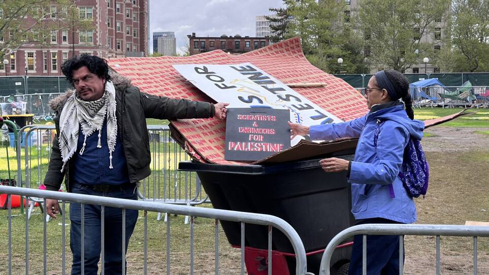 A person in a black and white keffiyeh gestures toward a sign reading scientists and engineers for Palestine in a trash barrel.