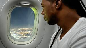 A man with a few hanging dreadlocks looks out the window of a plane.