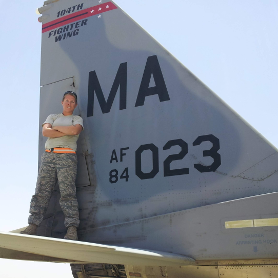 A woman in camo pants stands on a jet with her arms crossed.