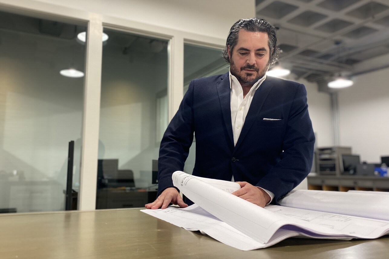 A man wearing a suit turns the page of a large booklet showing apartment floor plans. He is in what appears to be an otherwise empty large office.