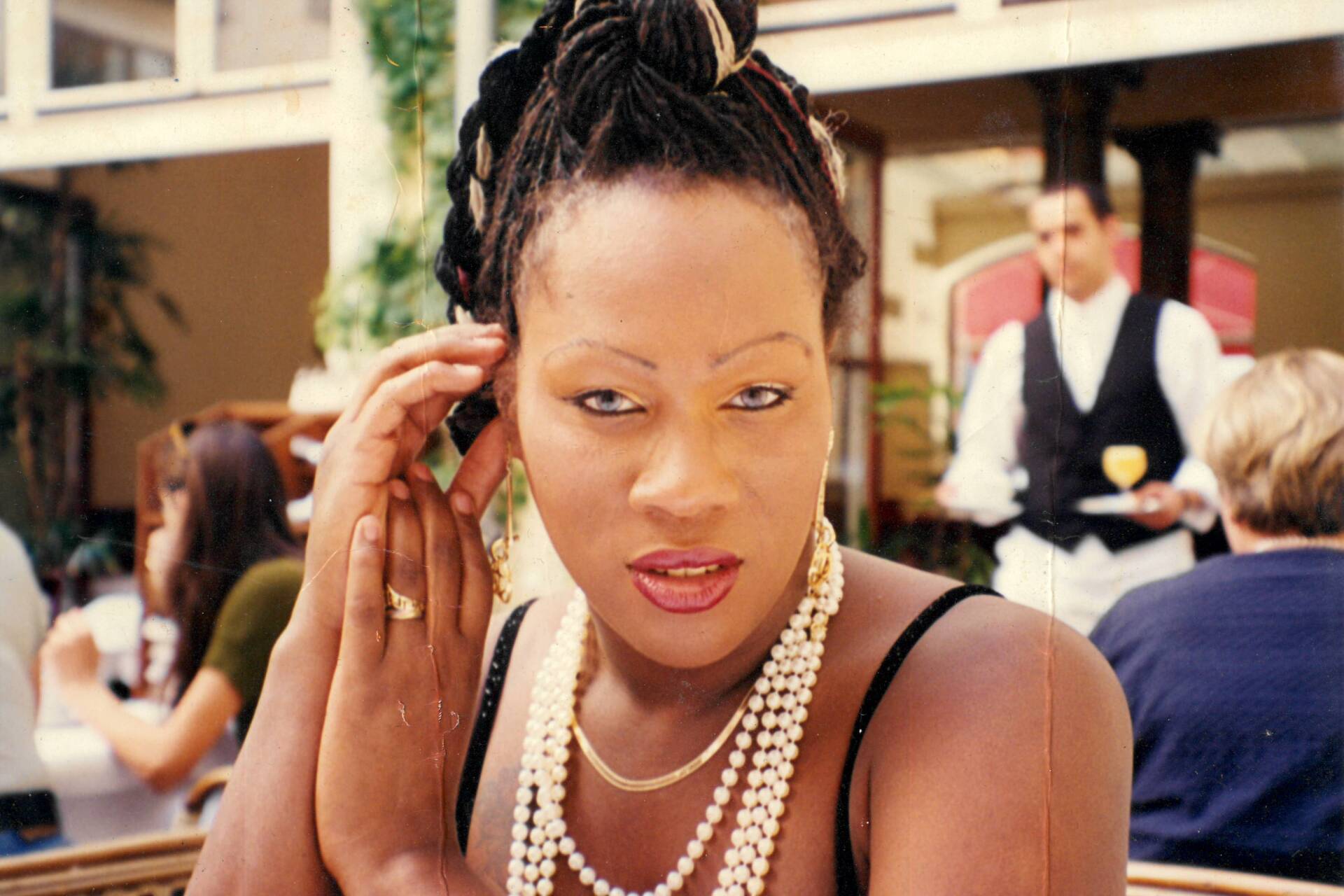 A woman, decked out in pearl necklaces and gold bracelets, holds her hands by her face as she sits at an outdoor restaurant.
