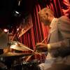 Joe Farnsworth sits at a drum kit in front of a red curtain.