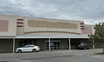 Outside an empty strip mall store with the sign above the doorway plastered over so the sign is blank. A sky heavy with dark clouds looms overhead. 