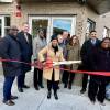 9 men and women stand outside a building behind a ceremonial red tape and one woman holds a giant pair of scissors ready to cut the tape.