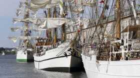 Three tall ships with multiple white sails and lots of ropes are docked in the water off a pier.