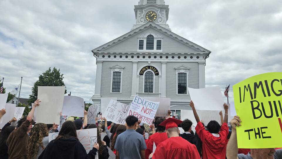 Protesters, including some in red graduation gowns, hold signs outside a white building with a clock tower.