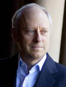 an older white man wearing a blue shirt and blazer gazes out a window with a contemplative look