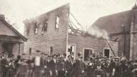 Black and white image of an assembly of men and boys in suits standing smiling in front of a wooden building that has been burned out by fire