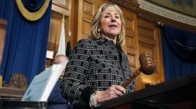 A woman in a business-y black-and-white patterned jacket holds a gavel as she stands behind a speaker's podium in the Massachusetts Statehouse.