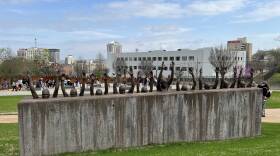 A vertical slab of concrete has a series of bronze renderings of people, rising out of the concrete with arms raised over their heads We see only their arms and heads. The image suggests people raising their hands, as if police officers were commanding them to have their "hands up." This is memorial on the grounds of the Memorial for Peace and Justice in Montgomery, Alabama.