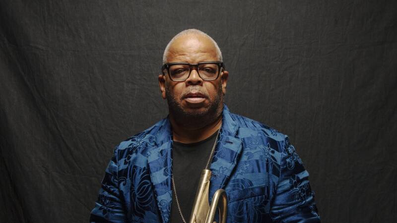 Terence Blanchard wears a blue velvet jacket and holds his trumpet against a black backdrop, a slight smile on his face.