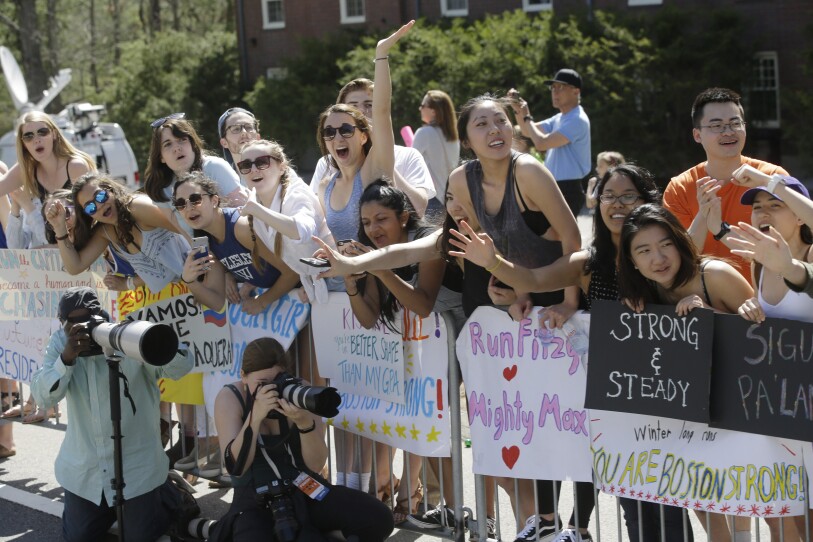 A group of mostly young women cheer excitedly over the rails of barricades with large posters to cheer on runners.