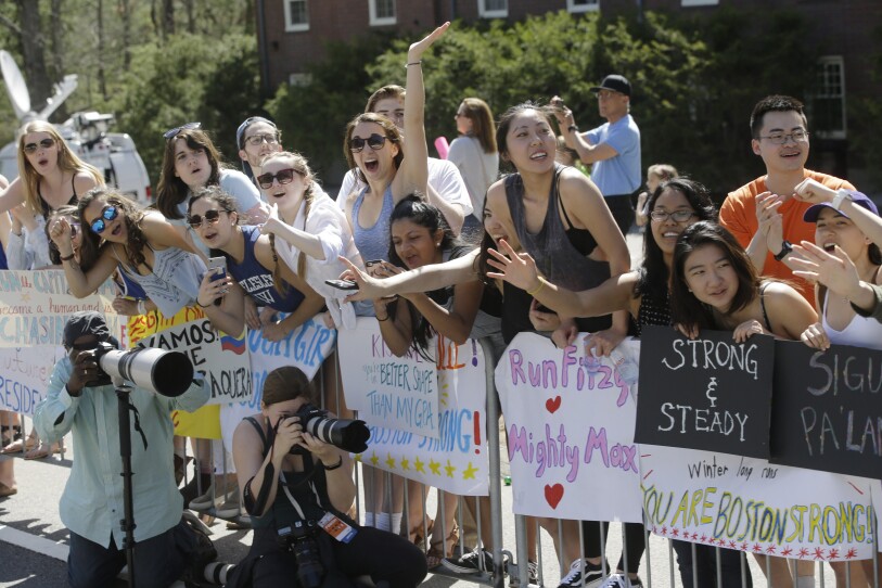 A group of mostly young women cheer excitedly over the rails of barricades with large posters to cheer on runners. 